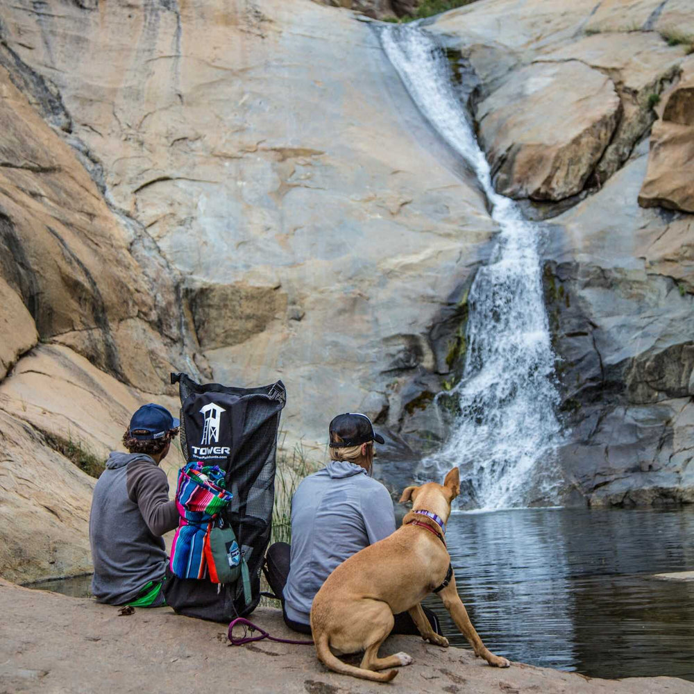 Couple with a dog holding their loaded up iSUP backpack from Tower while sitting near a waterfall and pond