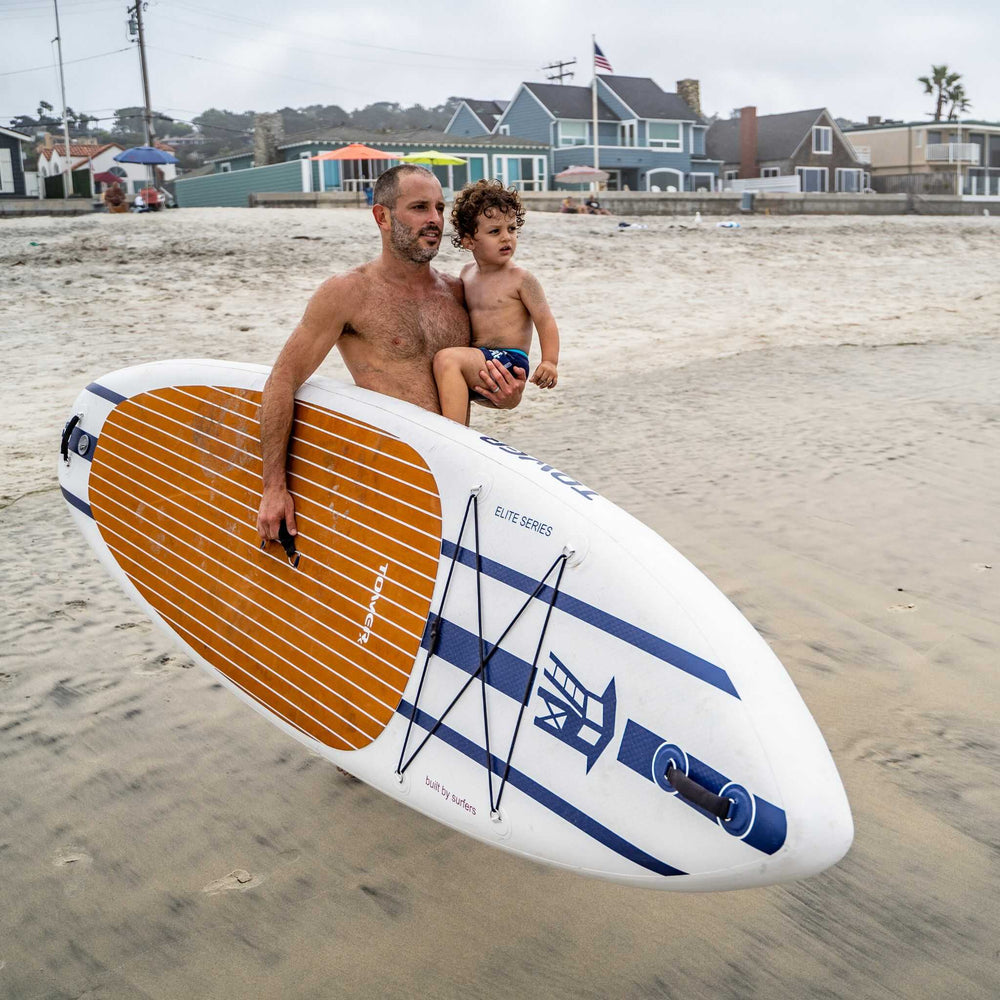 Man standing holding X-Class paddle board in one arm, with a child in the other