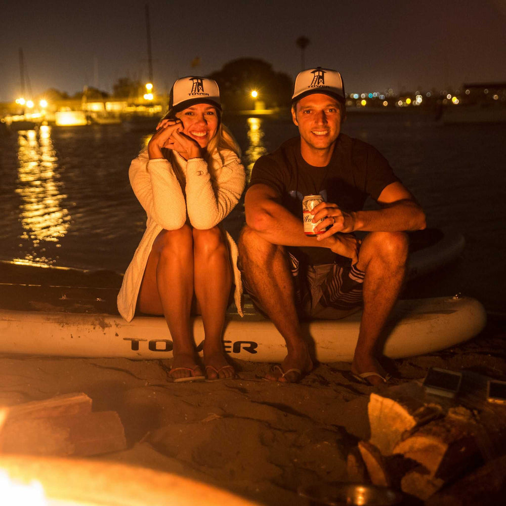 Man and woman sitting by a bonfire sitting on a Tower paddle board while wearing Tower trucker hats